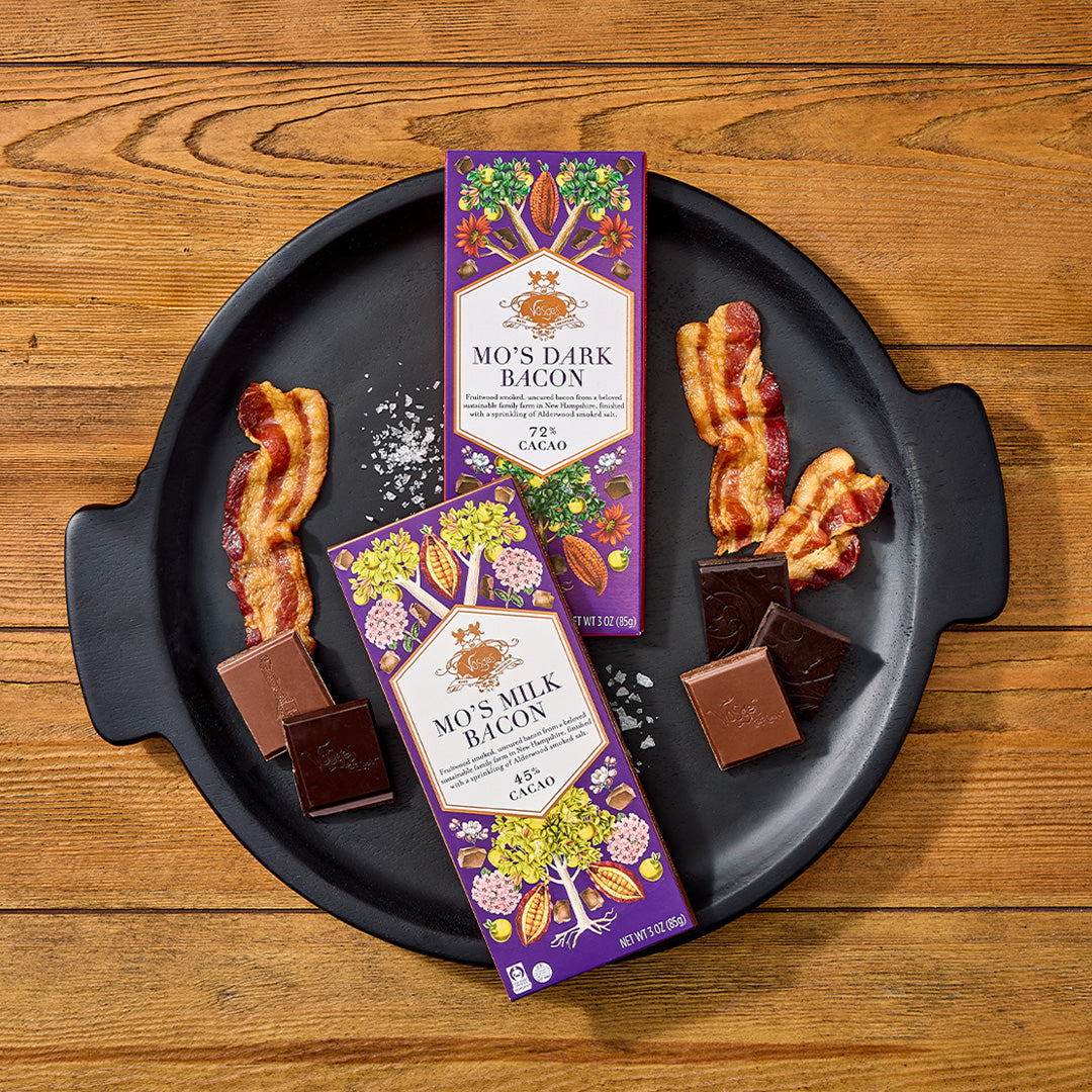 Mo's Bacon chocolate bars on purple Vosges packaging arranged on a black plate with bacon and chocolate pieces