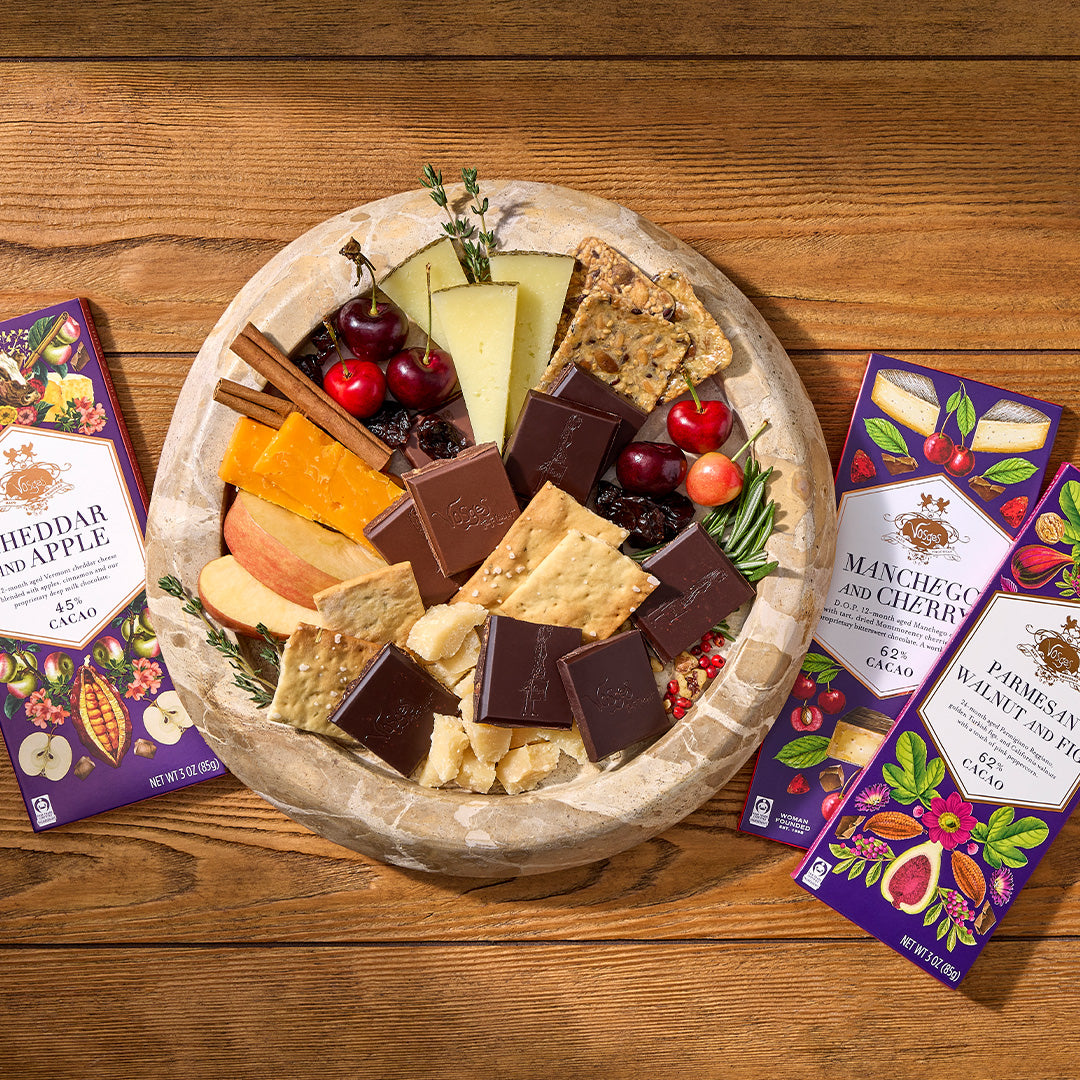 A round stone platter filled with assorted cheeses, crackers, dried fruit, and chocolate bars is arranged on a wooden table, with three purple packaged snack boxes placed beside it. The visible packages read Cheddar and Apple, Minimize and Cherry, and Minimize and Walnut Pie.