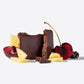 A chocolate bar styled with fruit pieces, including a bitten dark chocolate bar, pineapple chunks, a sliced red cherry, and dried cranberries, arranged on a clean white background.