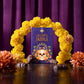 A boxed product labeled 'Chocolate Skulls' stands upright on a glossy dark surface, framed by a yellow flower garland and surrounded by chocolate pieces, against a purple curtain background.