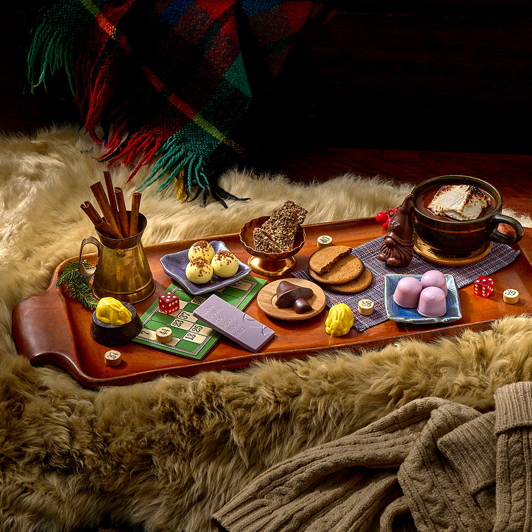 A cozy holiday-themed food and drink spread arranged on a wooden tray over a faux-fur surface, with a plaid blanket and knit sweater in the background. The tray holds a mug of hot chocolate, pastries, cookies, chocolates, dice, and small decorative items.