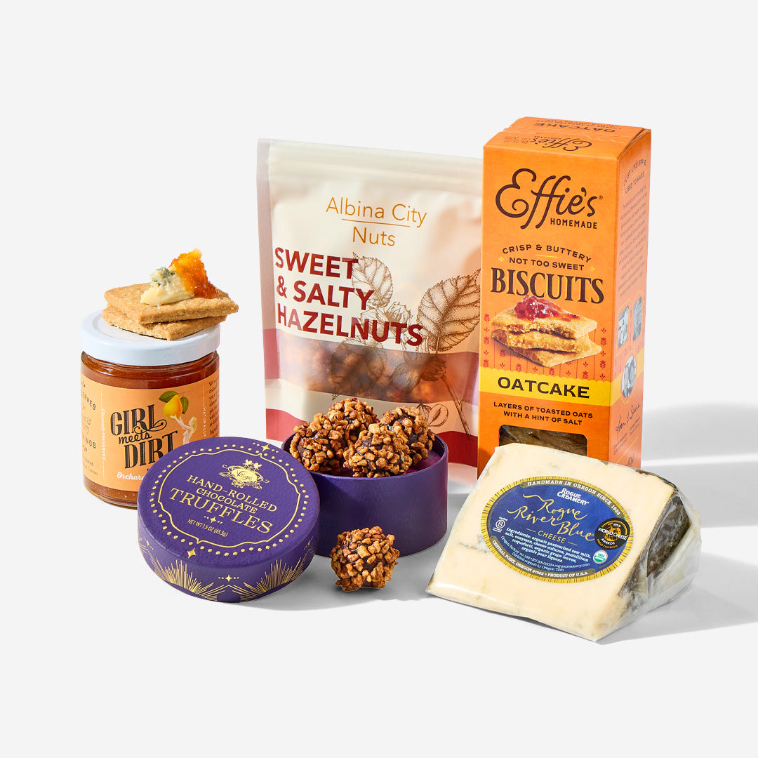 A curated gourmet snack and cheese assortment on a clean white background, featuring a jar of Girl and Dirt honeycomb crackers, a bag of Albina City Sweet & Salty Hazelnuts, a box of Effie's Oatcake Biscuits, a tin of Handcrafted Truffles, and a wedge of Roque Asnado Blue Cheese.