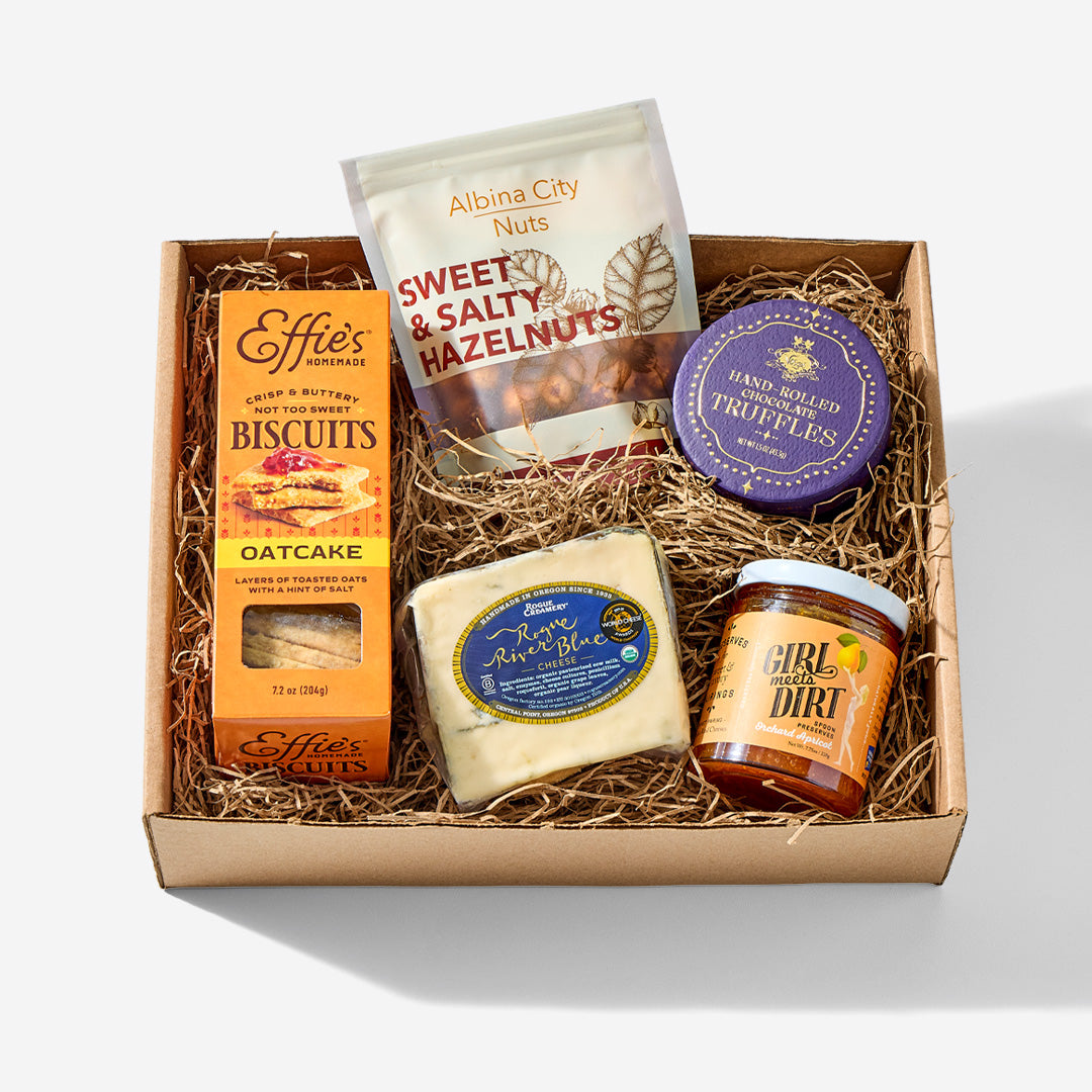 A curated gourmet gift box from Effie's and Albina City containing a box of oatcake biscuits, sweet and salty hazelnuts, truffles, a cheese wheel, and a jar of Gibrli 'Dirty' honey, arranged in a cardboard box with shredded paper.