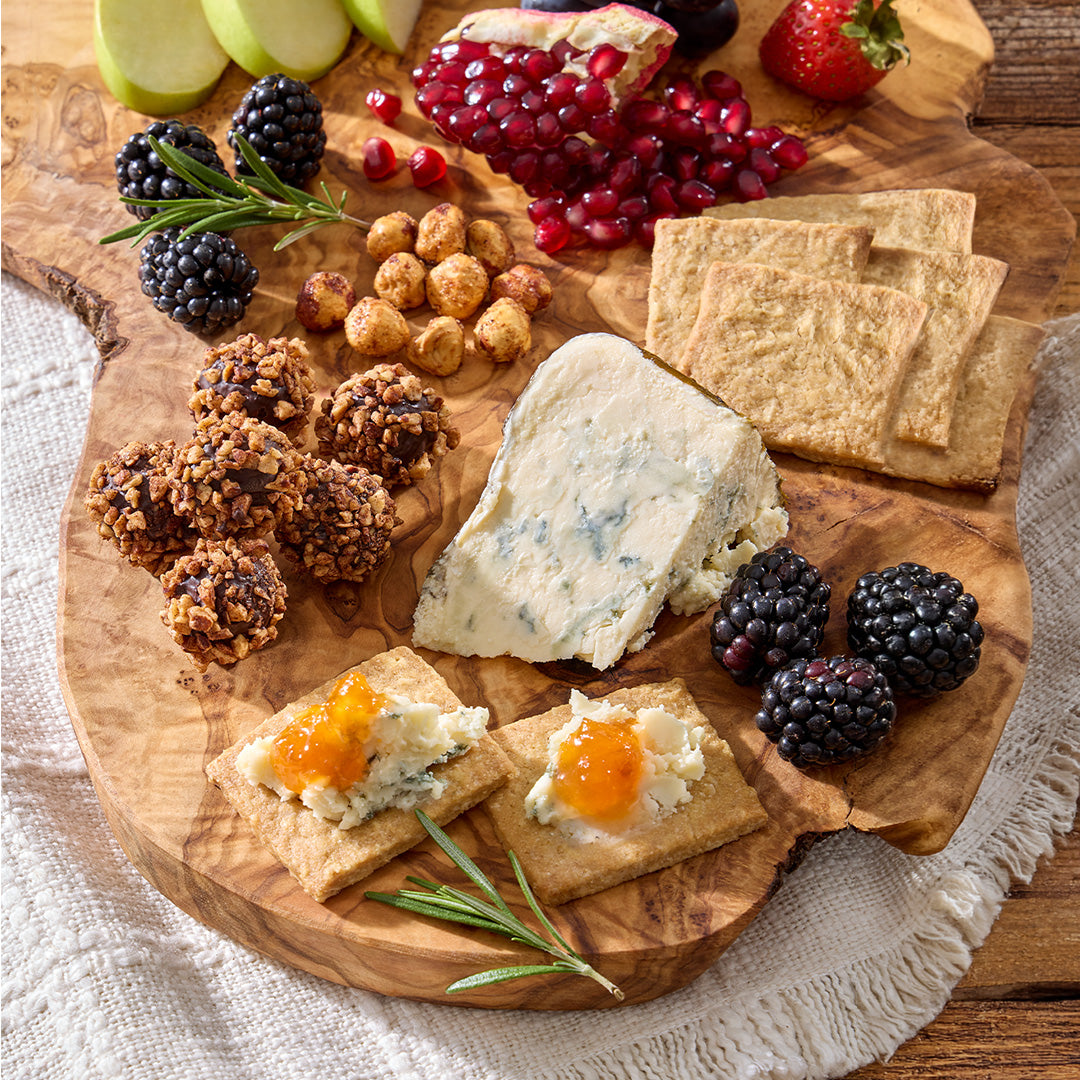 An artisanal cheese and charcuterie board arranged on a rustic wooden serving platter. The board includes a large wedge of blue cheese, crackers topped with white cheese and orange jam, blackberries, pomegranate seeds, nuts, and rosemary sprigs, set on a white textured cloth over a wooden table.