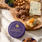 A round purple tin labeled 'HAND-ROLLED CHOCOLATE TRUFFLES' sits on a rustic wooden board styled with truffle balls, blue cheese, blackberries, and a cracker with orange spread, on a cream linen cloth.
