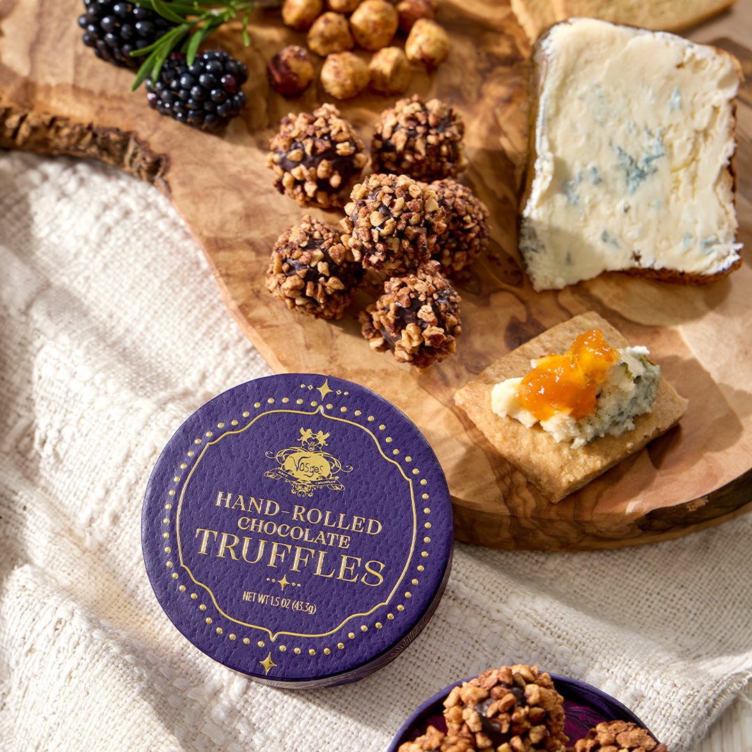 A round purple tin labeled 'HAND-ROLLED CHOCOLATE TRUFFLES' sits on a rustic wooden board styled with truffle balls, blue cheese, blackberries, and a cracker with orange spread, on a cream linen cloth.