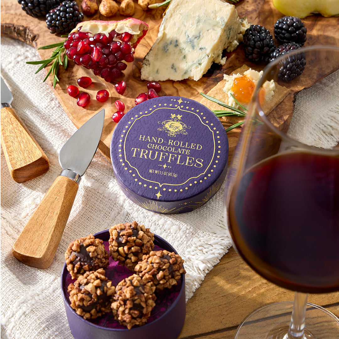 A round purple tin of hand-rolled chocolate truffles sits on a wooden charcuterie board surrounded by cheese, blackberries, pomegranate, rosemary, and a glass of red wine. A smaller purple container in front holds several truffles, with wooden-handled knives nearby.