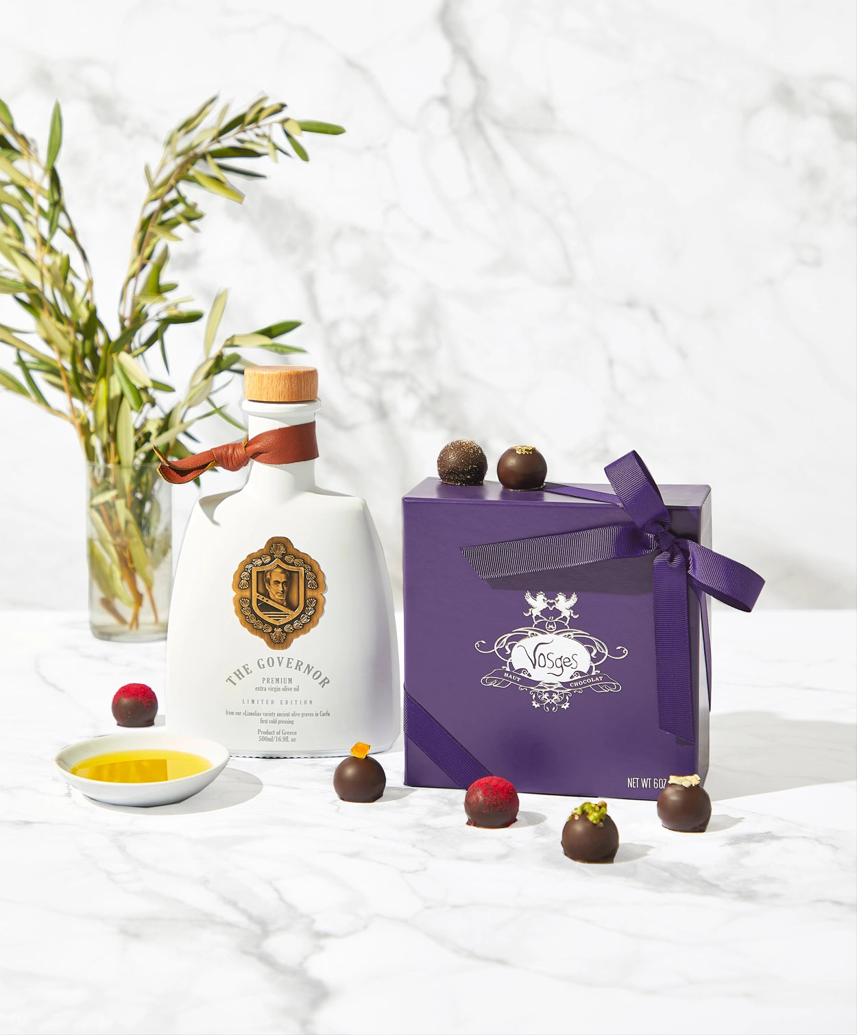 A bottle of Kyoord olive oil tied with a leather strap sits beside a purple candy box and a dish of oil surrounded by brightly decorated chocolate truffles on a marble background.