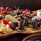 Three tall dark chocolate cone truffles on a rustic wooden board with cheese, fruit, and chocolates