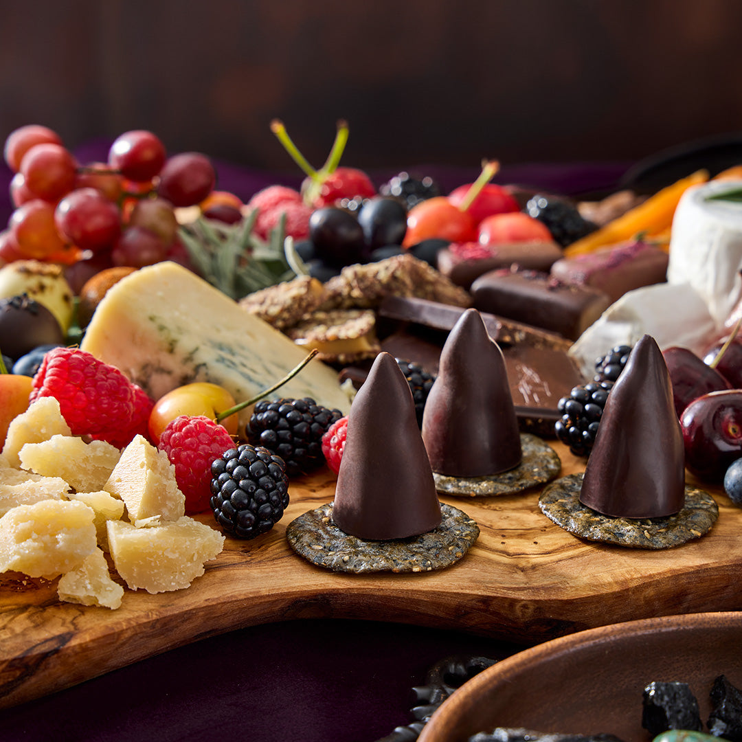 Three tall dark chocolate cone truffles on a rustic wooden board with cheese, fruit, and chocolates