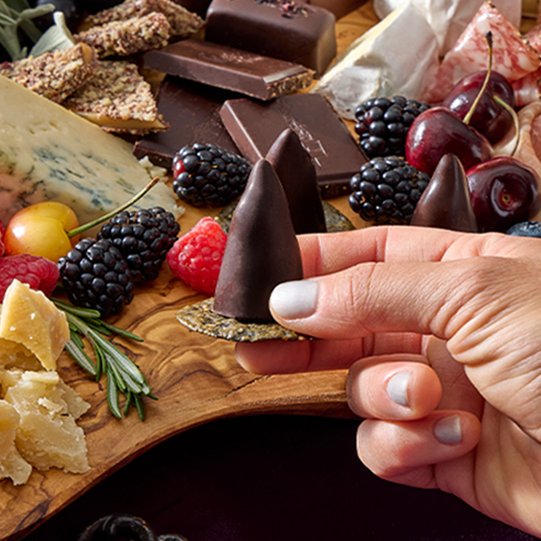 Hand holding a cone shaped chocolate from Spellbound Chocolate Menagerie over a cheese and fruit board