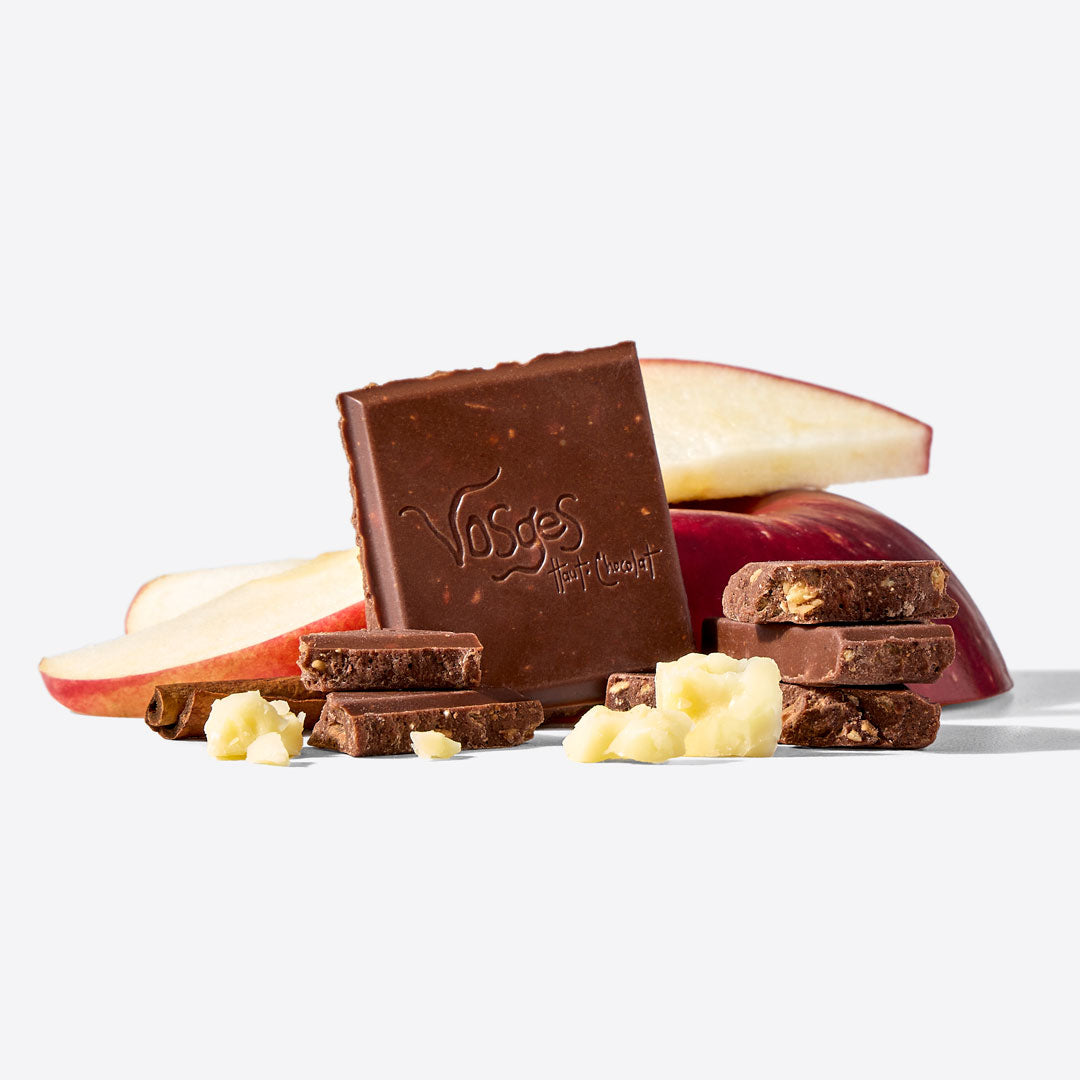 A square bar of Vosges dark chocolate with pieces broken and scattered in front, styled with red apple slices and white chocolate chunks on a clean white background.